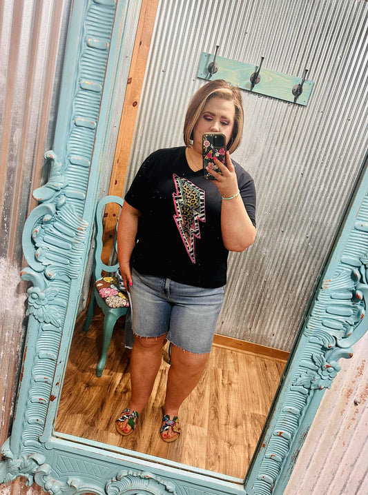Woman wearing light wash mid-length denim shorts with a raw hem, standing in front of a mirror inside a boutique.