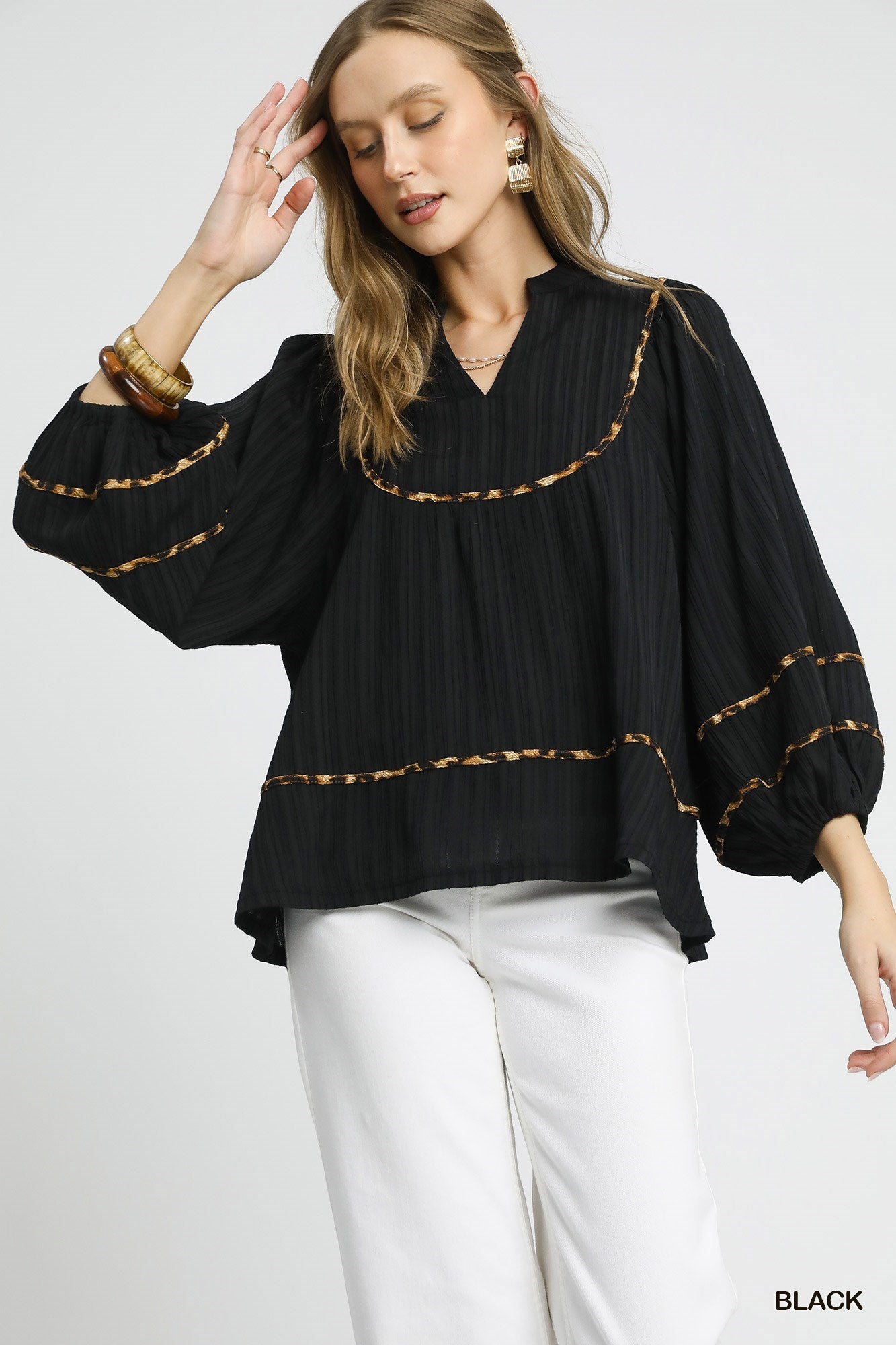 Woman wearing a flowy black blouse with golden embroidery and balloon sleeves, paired with white pants.