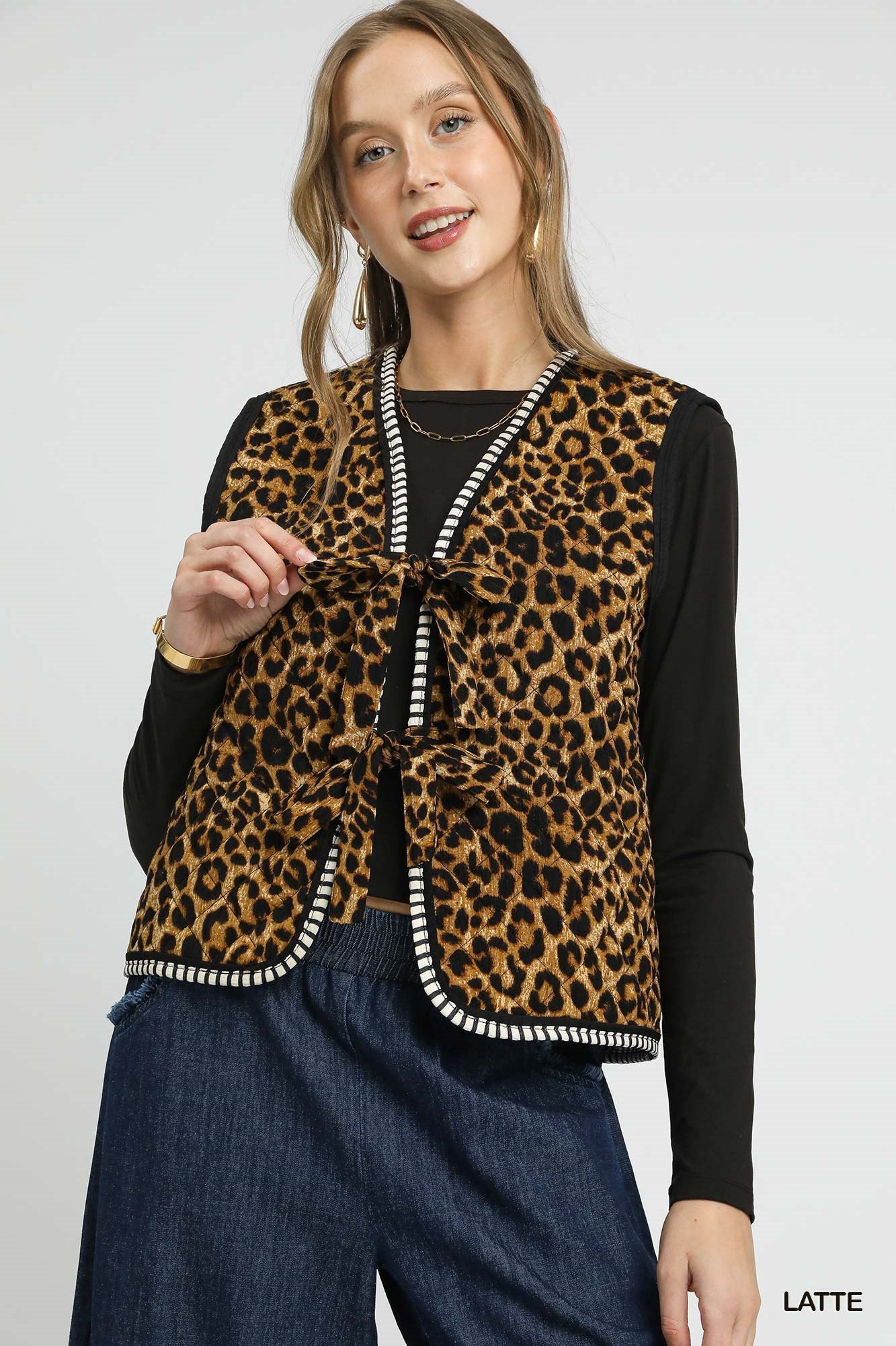 Woman wearing a sleeveless leopard print vest with bow tie front and contrast striped trim over a black long sleeve top.