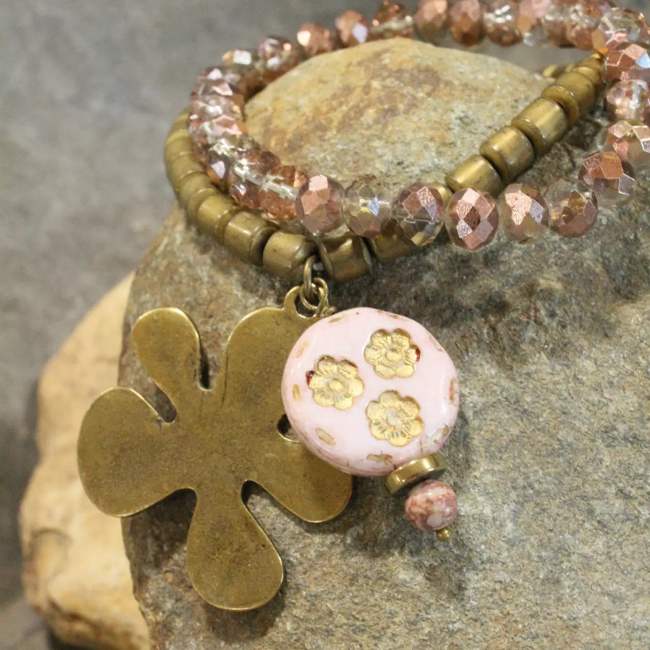 Blush and bronze beaded bracelet set featuring a clover charm and pink floral ceramic bead, displayed on a rock surface.