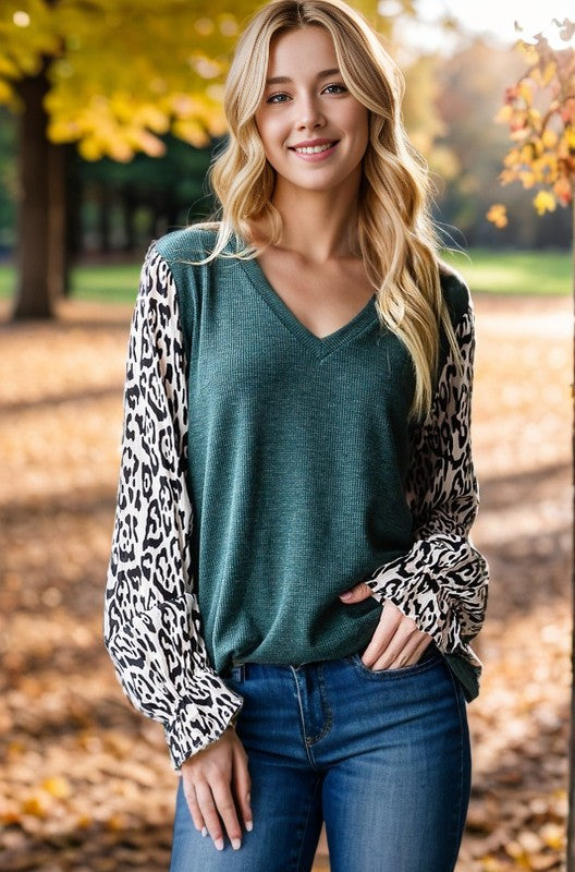 Model wearing a teal knit v-neck top with leopard print balloon sleeves and jeans in a fall outdoor setting.


