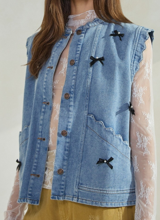 Woman wearing acid washed denim vest with bow and pearl accents over a lace blouse.