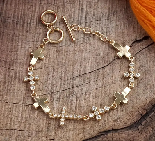 Gold cross bracelet with toggle closure and rhinestone accents displayed on wood background.